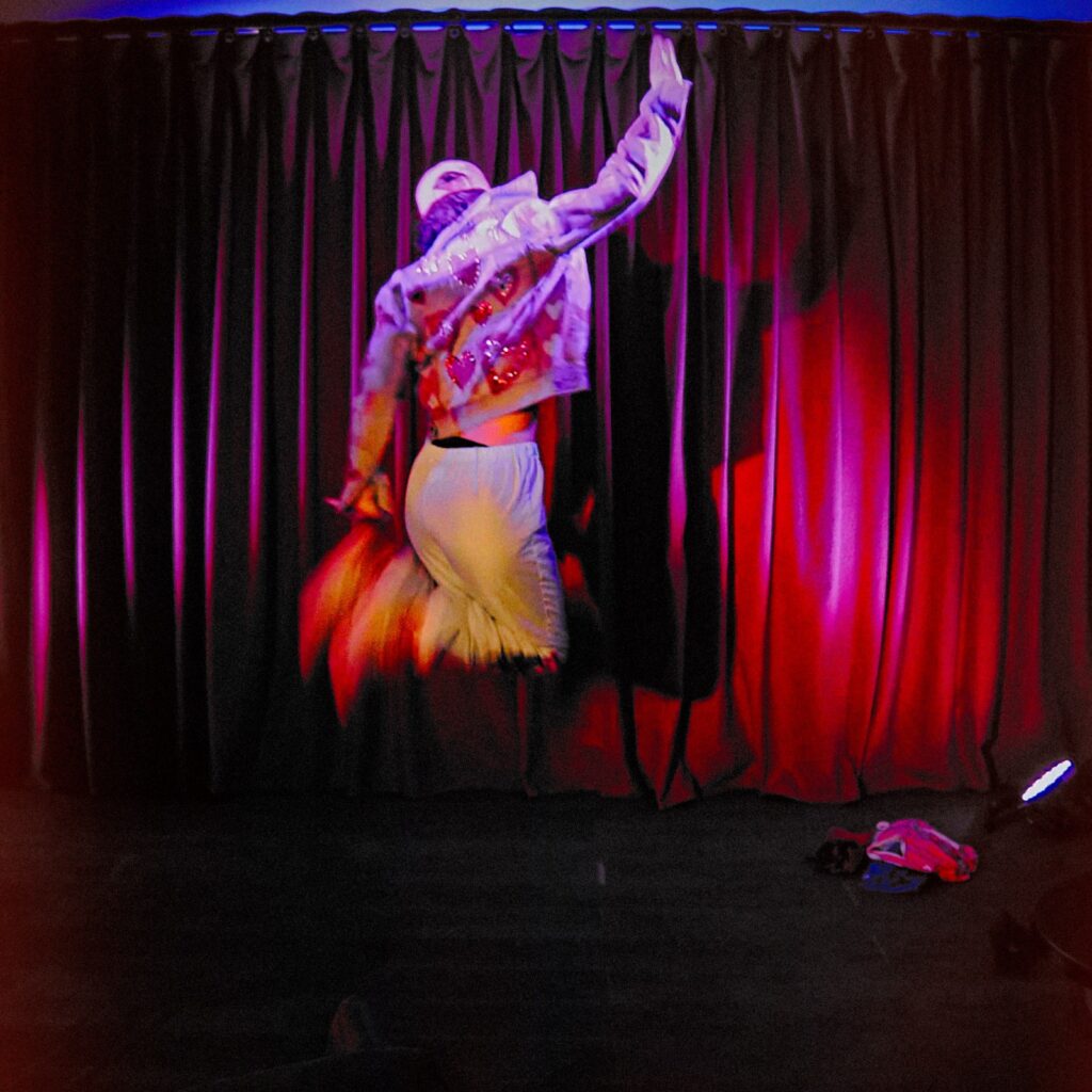 A person in a cap, heart-patterned jacket, athletic bottoms and trainers faces away from the camera, mid-jump, as they touch their feet with one hand and raise the other to the sky.