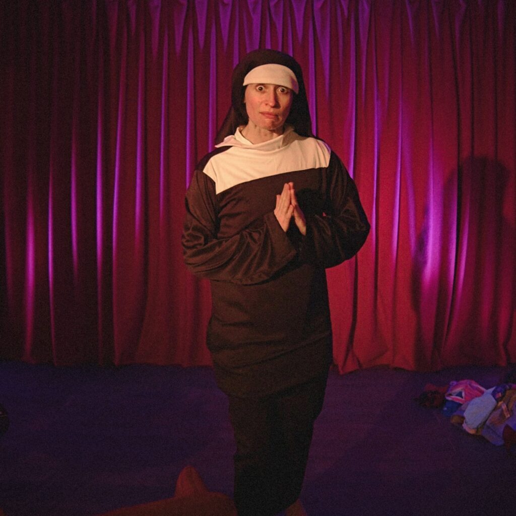 A woman in a nun habit stands before a red curtain, hands folded as if in prayer.